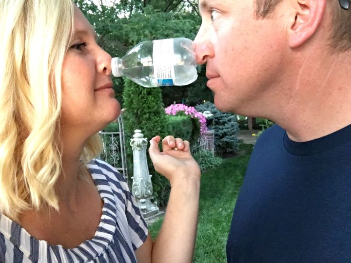 A couple facing each other and holding a small bottle of water up by pressing their noses against the bottle from either side.